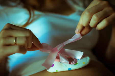 A pregnant woman holding white shoe and pink bow ribbon on pregnancy belly. Waiting for daughter.