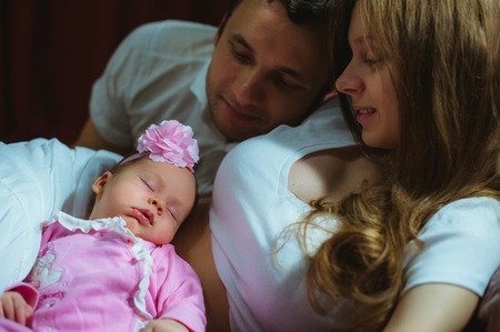 Image of young caucasian family indoor. Father, mother and cute little girl. Dad, mum and newborn baby in pink suit.の写真素材