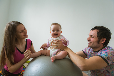 Child jumps on ball with help of parents. Image of young dad and mama with cute little daughter on floor at home. Father, mother and baby girl indoor.の写真素材