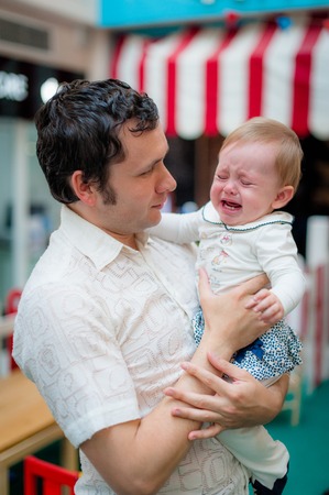 Unhappy child in the hands of daddy. Image of young dad with cute little daughter in his arms. Father and baby girl child in cafe.の写真素材