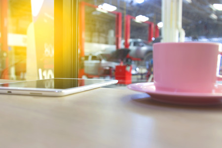 Wood table top and tablet, coffee cup with Car repair service centre interior blurred background. For create montage auto product display.の写真素材