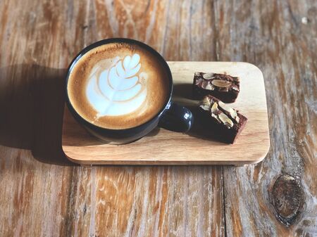 Top view Delicious Breakfast;  Latte art coffee in Black cup and Brownie topped with almond sliced in wooden tray on the vintage wooden table.の写真素材
