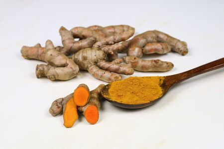Turmeric powder in a wooden spoon and fresh turmeric roots isolated on a white background is an...