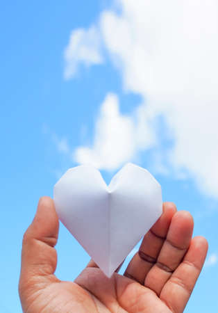 Hand holding a white paper heart on the sky background.の写真素材