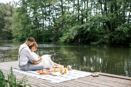 Happy couple have a rest in the park near the lake on a picnic. A couple sits on a plaid blanket on a picnic near a meal, hug and relax. Summer picnicの写真素材