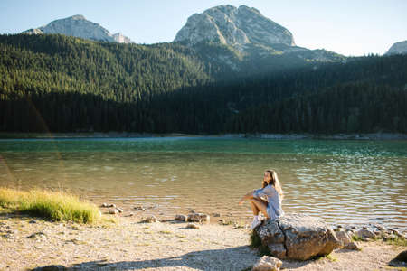 Happy woman tourist sits on a stone near the lake, rests and breathes the mountain air. View of the lake, forest and mountains.の写真素材