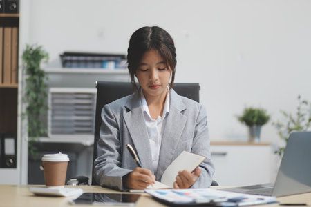 Successful Asian businesswoman smiling using laptop computer and holding coffee cup at office. Confident Asia businesswoman sitting happily in the office.の写真素材