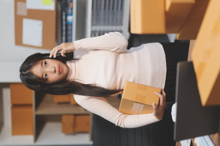 Startup SME small business entrepreneur of freelance Asian woman wearing apron using laptop and box to receive and review orders online to prepare to pack sell to customers, online sme business ideas.の写真素材