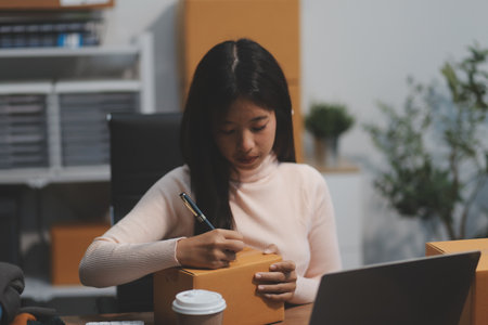 Startup SME small business entrepreneur of freelance Asian woman wearing apron using laptop and box to receive and review orders online to prepare to pack sell to customers, online sme business ideas.の写真素材