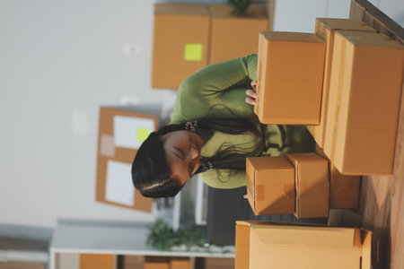 Startup SME small business entrepreneur of freelance Asian woman wearing apron using laptop and box to receive and review orders online to prepare to pack sell to customers, online sme business ideas.の写真素材