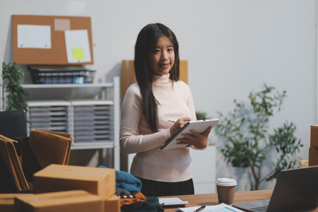 Startup SME small business entrepreneur of freelance Asian woman wearing apron using laptop and box to receive and review orders online to prepare to pack sell to customers, online sme business ideas.の写真素材
