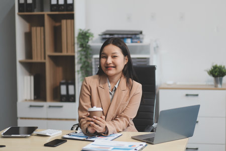Attractive Asian secretary or businesswoman holding hot coffee mug sitting on laptop in office.の写真素材