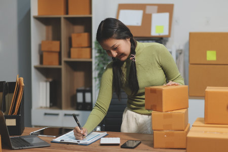 Startup SME small business entrepreneur of freelance Asian woman wearing apron using laptop and box to receive and review orders online to prepare to pack sell to customers, online sme business ideas.の写真素材