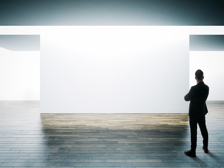 Businessman stands opposite big white wall in museum interior with wooden floor.の写真素材