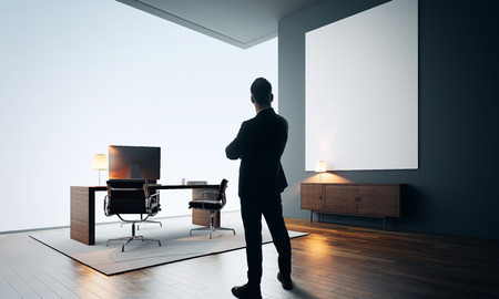 Businessman stands in contemporary office interior. Workspace in loft with generic design computer, wooden floor and one empty canvas.の写真素材