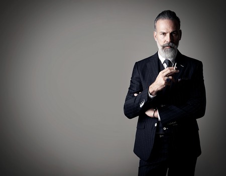 Portrait of smoking gentleman wearing trendy suit and stands against the empty wall.