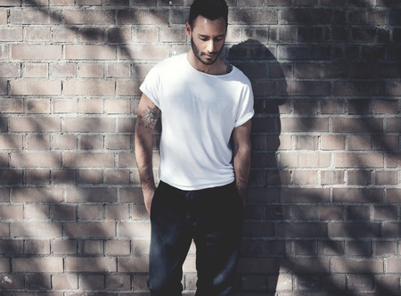 Bearded man with tattoo wearing blank white tshirt and black jeans. Bricks wall background....