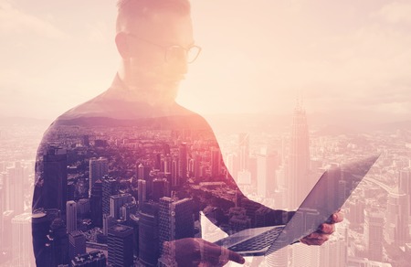 Double exposure businessman wearing black shirt and glasses.Banker holding contemporary notebook hands.Using work online banking system. Modern city sunset background.