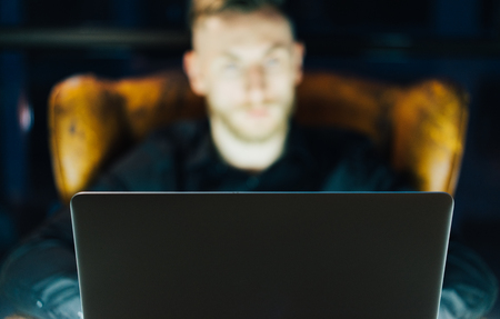 Photo young banker working on modern loft office. Man sitting in vintage chair at night. Using contemporary laptop, blurred background. Soft light,shadows.の写真素材