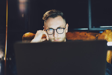 Photo young manager working on modern loft office. Man sitting in vintage chair at night. Using contemporary laptop, blurred background. Soft light,shadows.の写真素材
