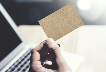 Closeup Photo Man Showing Blank Craft Business Card and Using  Modern Laptop.Wood table Blurred Background. Mockup Ready for Private Information.Sunlight Reflections Screen Gadget.Horizontal mock upの写真素材