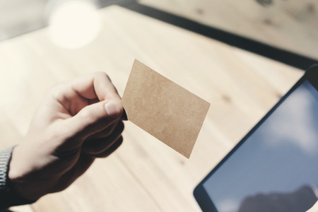 Closeup Man Showing Blank Craft Business Card, Holding Hand Modern Digital Tablet.Wood table Blurred Background.Mockup Ready  Private Information.Sunlight Reflections Screen Gadget.Horizontal mock upの写真素材