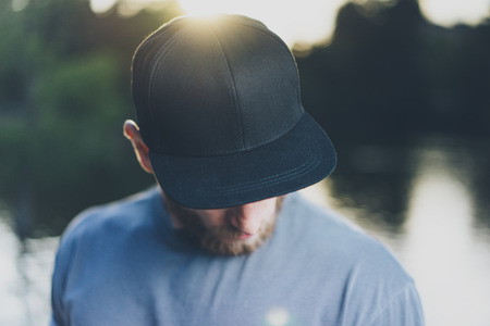 Photo Bearded Young Man Wearing Black Blank Cap. Green City Park Lake Background and Sunset effect....