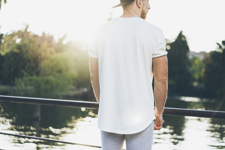 Picture Bearded Muscular Man Wearing White Blank t-shirt, snapback cap and shorts in sunset time. Relaxing time near the lake. Green City Garden Park Sunset Background. Back view. Horizontal Mockup.の写真素材