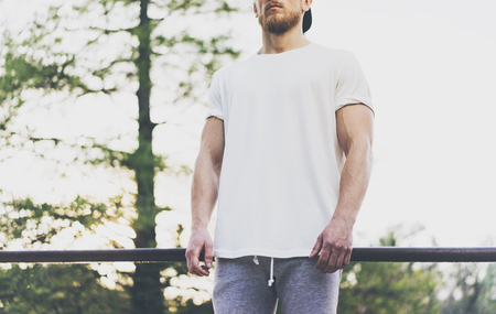 Photo Bearded Muscular Man Wearing White Blank t-shirt, snapback cap and shorts in summer vacation. Relaxing time near the lake. Green City Garden Park Sunset Background.Front view. Horizontal Mockup.の写真素材