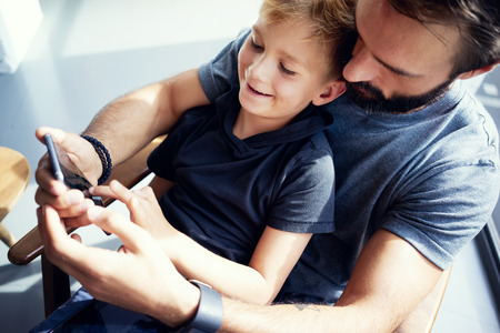 Closeup of young boy sitting with father and using smartphone in modern sunny place. Horizontal, blurred backgroundの写真素材