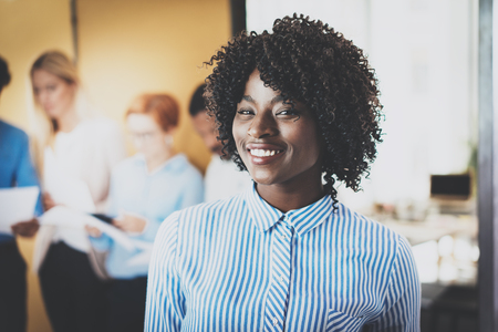 Portrait of pretty african american business woman with afro smiling at the camera.Coworking team on background in modern office. Horizontal,blurredの写真素材