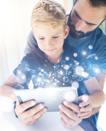 Father and his little son playing together on mobile computer, resting indoor.Bearded man with young boy using tablet PC in sunny house.Childhood dreams icons concept.Vertical, blurred backgroundの写真素材
