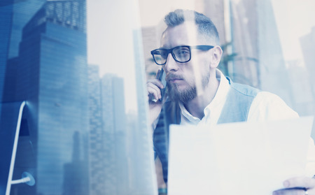 Bearded young businessman wearing white shirt,waistcoat and making call at the working place.Concept of successful business.Double exposure,skyscraper office building on blurred backgroundの写真素材
