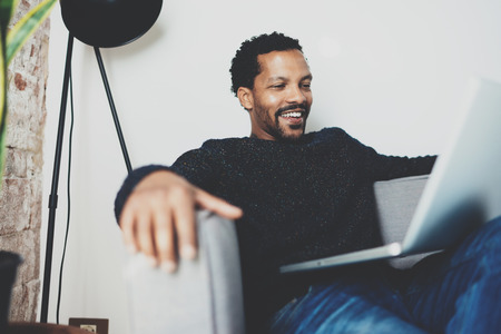 Attractive bearded African man laughing and holding laptop on knees while sitting sofa at his modern home office.Concept of young people enjoying mobile devices.Brick wall blurred backgroundの写真素材