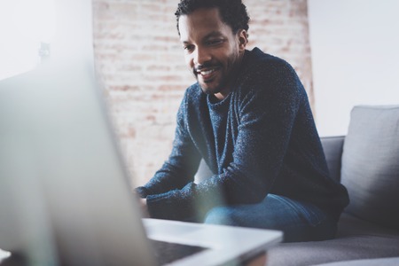 Cheerful bearded African guy working on laptop while sitting sofa at his modern office place.Concept of young people using mobile devices.Brick wall blurred background.Horizontalの写真素材