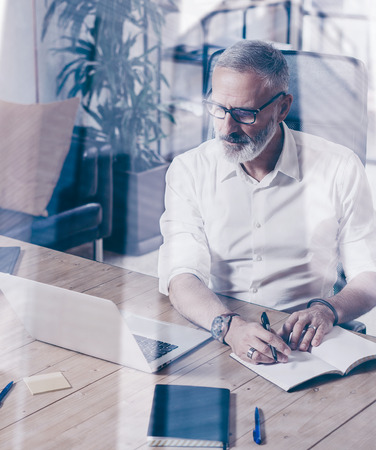 Concept of adult successful businessman wearing a classic glasses and working at the wood table in modern coworking place.Double exposure,skyscraper office building blurred background.Verticalの写真素材