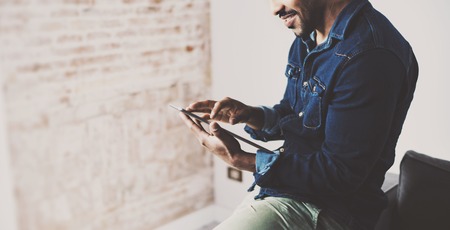 Attractive bearded African man using tablet at his empty room.Concept of young people enjoying mobile devices.Blurred brick wall on the background.Color filter.Wide.の写真素材