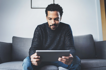 Attractive bearded African man using tablet while sitting on sofa in his modern office.Concept of young business people working at home.Blurred background.の写真素材