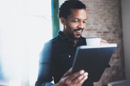 Closeup of young bearded African man using tablet while holding white cup coffee in hand at modern coworking office.Concept people working with mobile gadget.Blurred brick wall on the background.の写真素材