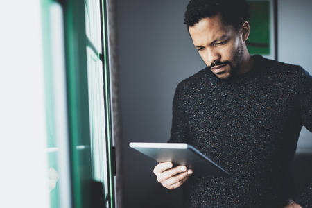 Selective focus.Pensive bearded African man using digital tablet while standing near the window in his modern apartment.Concept of young business people working at home.Blurred background.の写真素材