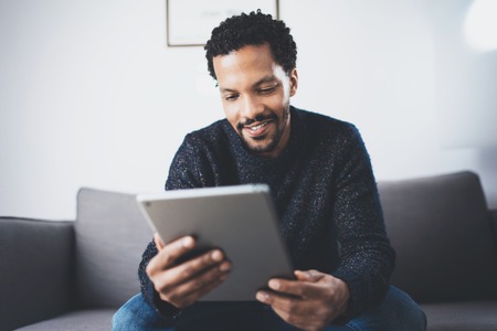 Selective focus.Attractive bearded African man reading news on digital tablet while sitting  sofa in...