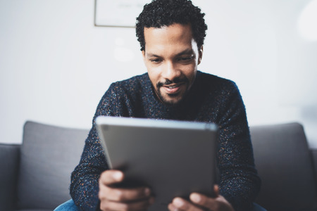 Selective focus.Closeup view of attractive bearded African man reading news on digital tablet while sitting sofa in his modern studio.Concept young business people working home.Blurred background.の写真素材