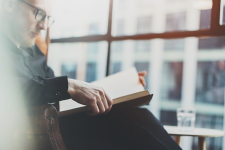Closeup portrait of pensive bearded businessman reading book while sitting in vintage chair.Young man relaxing at home.Selective focus on hands,blurred background.Horizontal, film effect.の写真素材