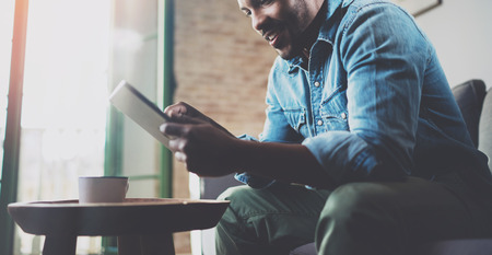Smiling bearded African man using tablet for reading morning news at home.Concept people working with mobile gadget.Cup of coffee on the wooden table.Blurred background,crop.の写真素材