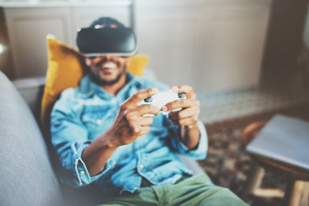 Smiling african man enjoying virtual reality glasses while relaxing on sofa.Young guy with vr headset or 3d spectacles and controller gamepad playing video game at home.Selective focus hand,blurred.の写真素材