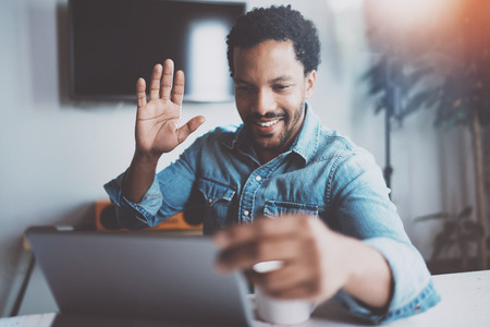 Smiling young African man making video conversation via digital tablet with business partners and greets them hand raised.Concept of happy coworking people.Blurred background,flare.の写真素材