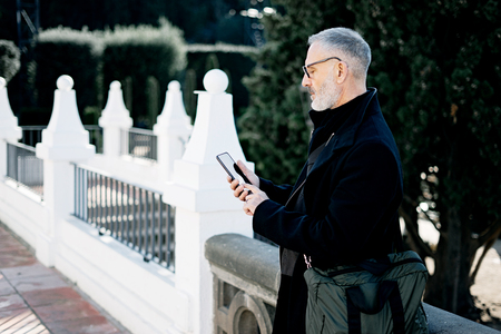 Attractive gray-haired businessman texting message on mobile phone while spending time in city park at sunny day.Horizontal,blurred background.の写真素材