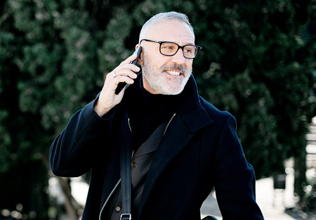 Portrait of smiling gray-haired man making call on his mobile phone while spending time in city park...