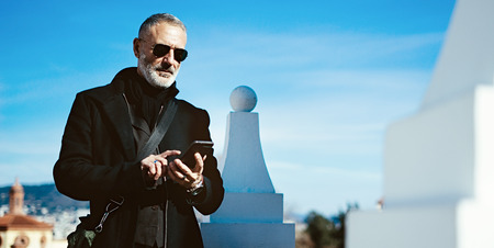 Pensive adult man wearing black aviator sunglasses and using smartphone while spending time in city park.Horizontal wide,blurred background.の写真素材