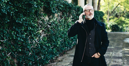 Portrait of successful middle age businessman talking on his smartphone while walking in city park.Horizontal wide,blurred background,film effect.の写真素材